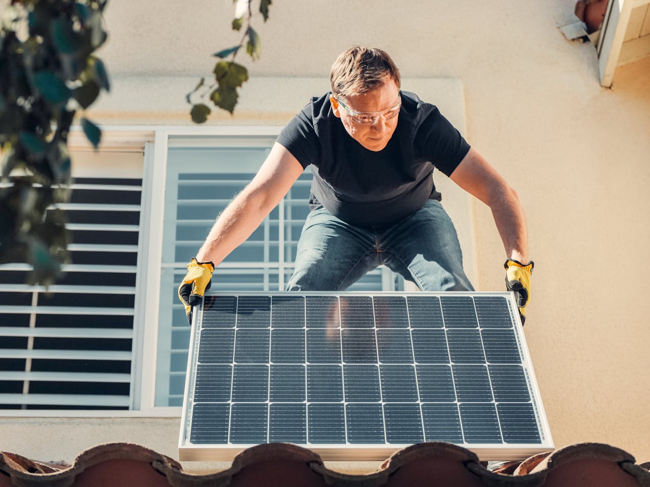 creative-02 A man in safety glasses installs a solar panel on a house roof, promoting renewable energy.