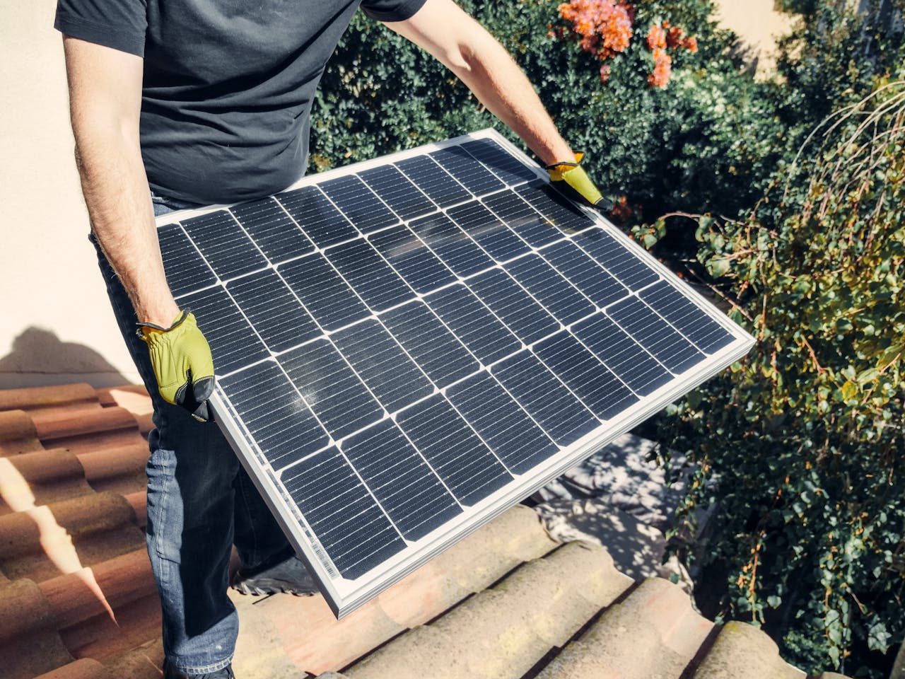 creative-03 A worker in gloves installs a solar panel on a tiled rooftop, promoting renewable energy.